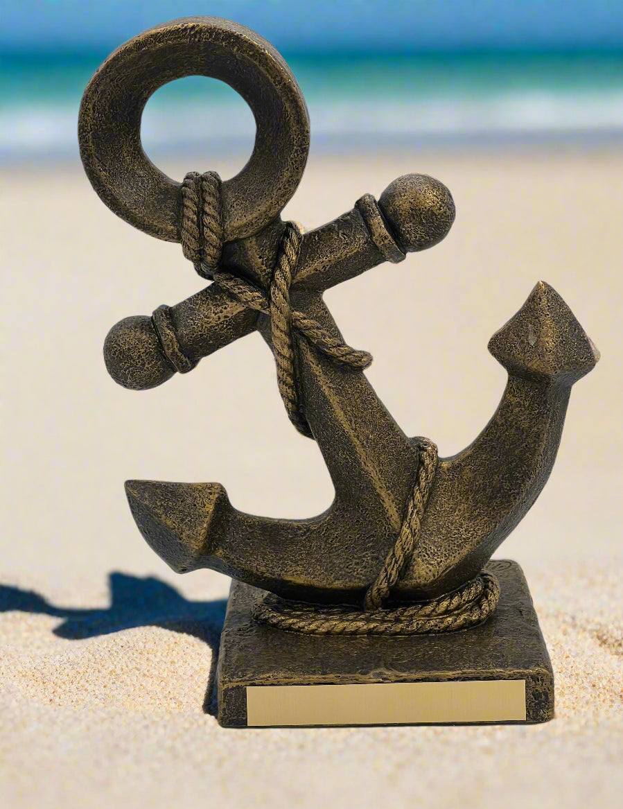 A bronze anchor sculpture sitting on a square base with a rope wrapped around its shank, placed on a sandy surface with a body of water and sky in the background.