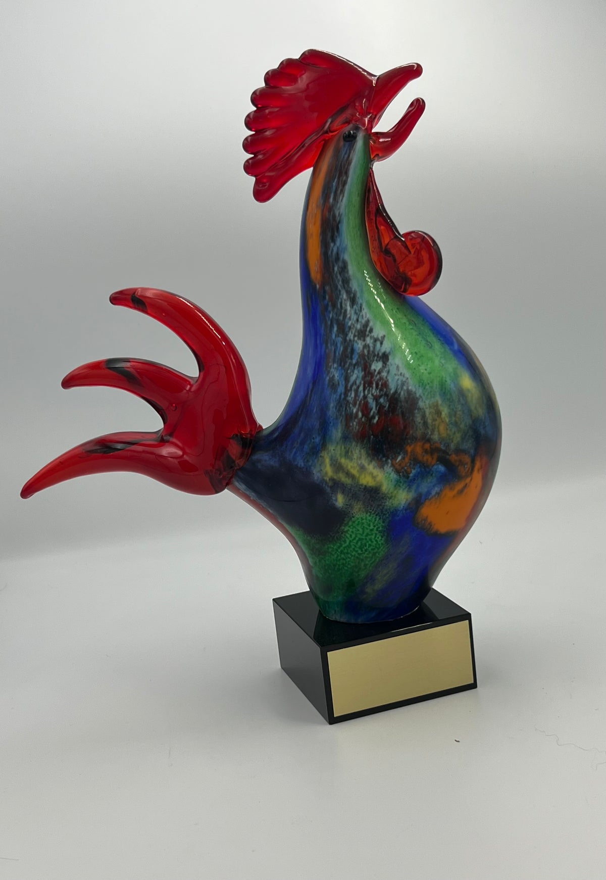 A colorful glass rooster sculpture on a black base, with a solid marble baseplate that has lettering.