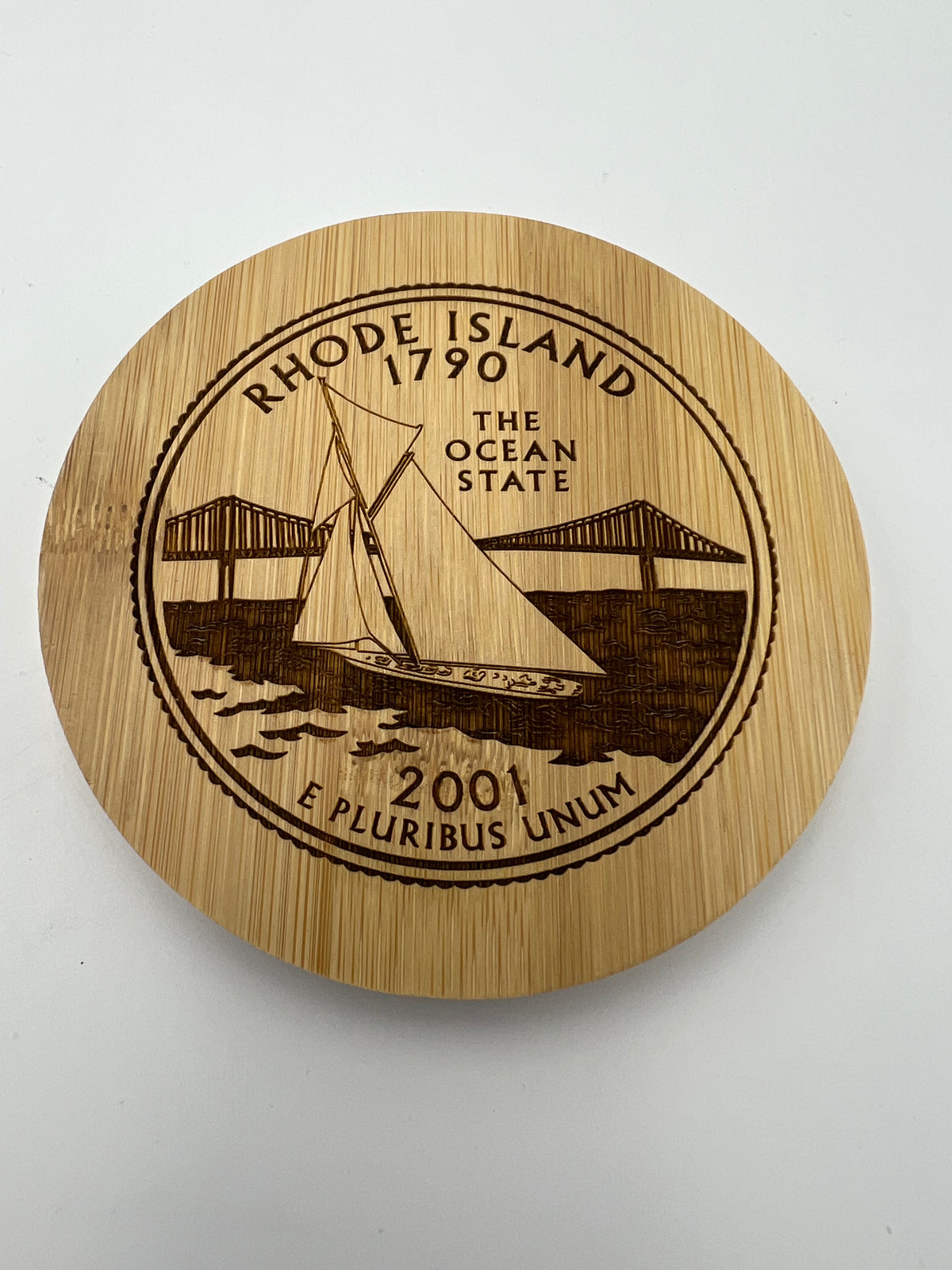 A bamboo coaster with an engraved image of a Rhode Island quarter, including the state seal and a sailboat.
